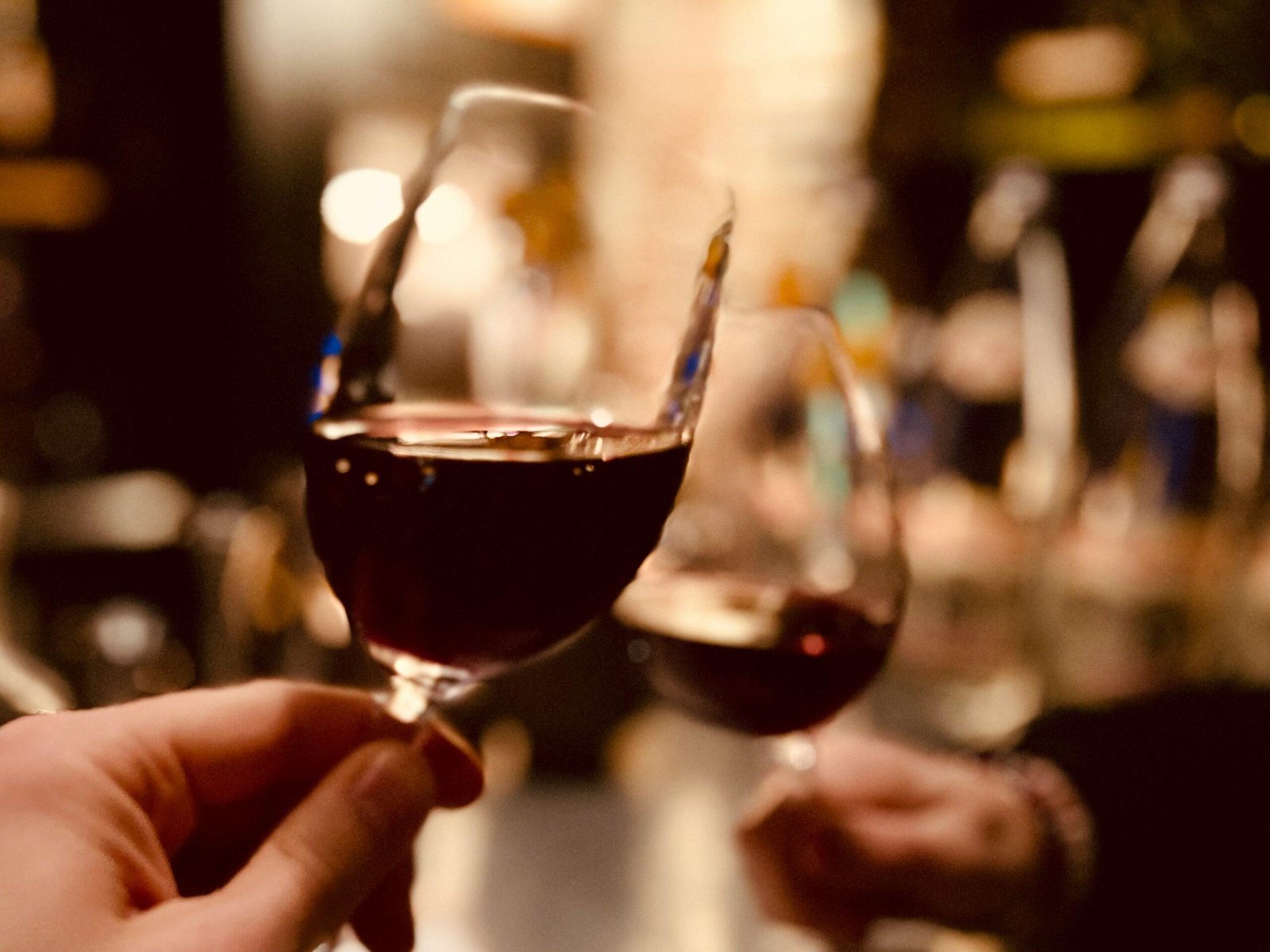 Two glasses of red wine being held and clinked for a toast in a dimly-lit setting, showcasing the perfect moment captured by skilled wine marketing. The blurred background enhances the warm and cozy atmosphere.