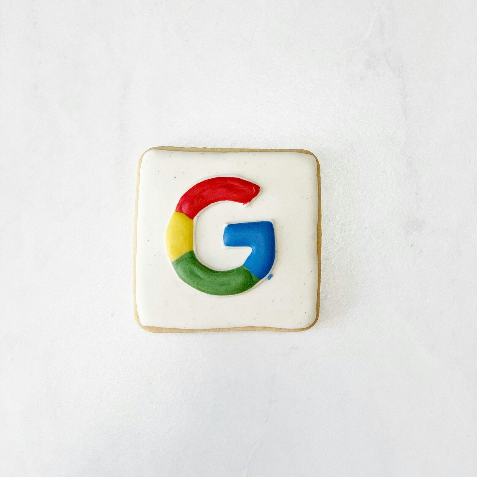 A square cookie decorated with the Google logo sits elegantly on a plain white surface. The top of the cookie is frosted white, showcasing a multicolored "G" with blue, red, yellow, and green segments—a sweet tribute to Google cookie deprecation delays and its impact for advertisers.