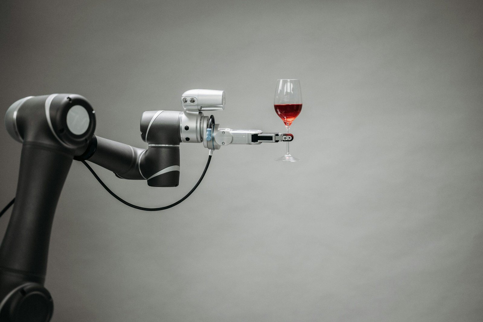 A robotic arm holds a glass of red wine against a plain gray background. The arm features sleek metallic components and a gripping mechanism. The wine glass is delicately balanced, showcasing precision and technology.